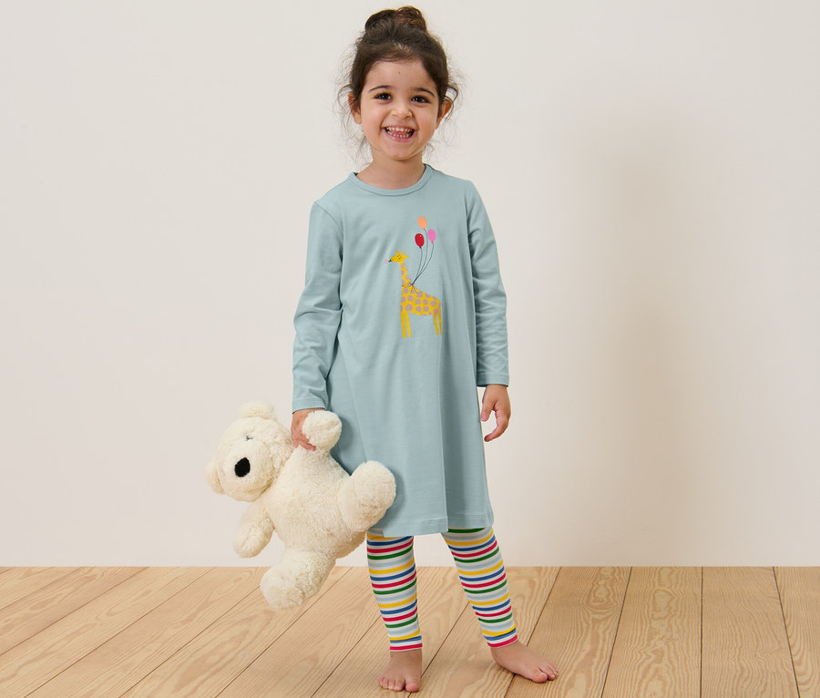 Lächelndes Mädchen hält Teddybär, trägt Kinder-Nachthemd mit Leggings.