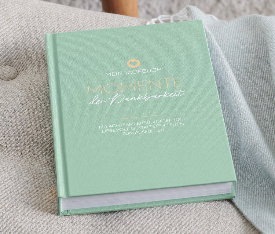 Ein grünes Tagebuch "Momente der Dankbarkeit" liegt auf einem beigen Sessel.