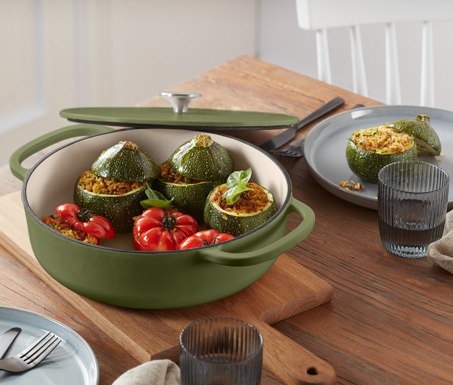 Eine dunkelgrüne Gusseisen-Bräter-Pfanne mit Deckel, gefüllt mit gefüllten Zucchini und Tomaten, steht auf einem Holzbrett auf einem Tisch.
