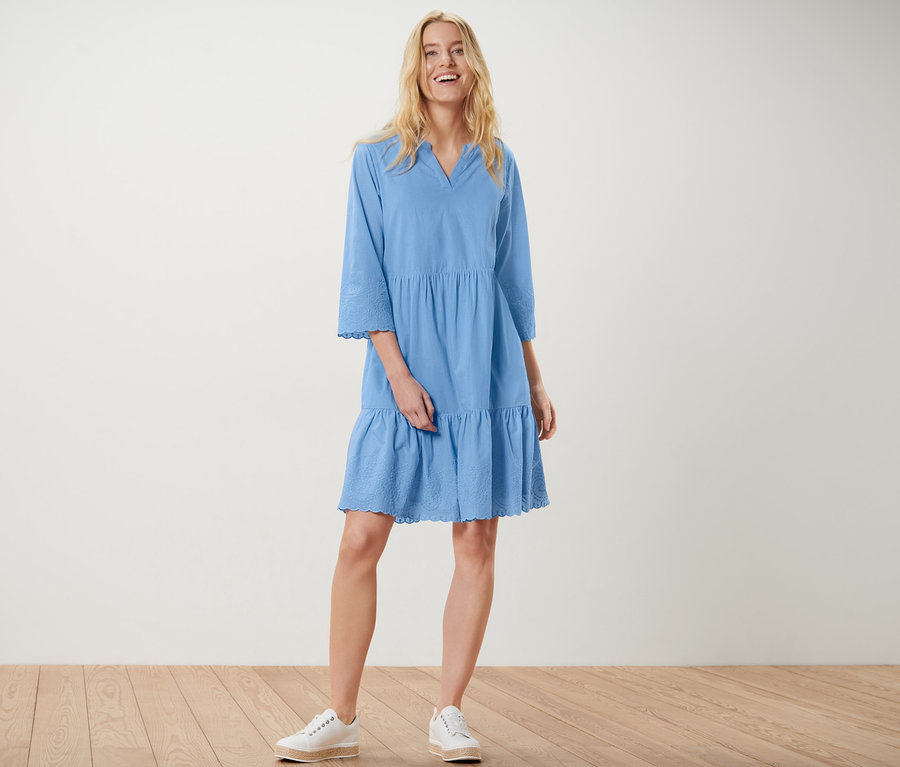 Eine blonde Frau steht und lächelt, sie trägt ein blaues Webkleid mit Stickerei und weiße Turnschuhe.