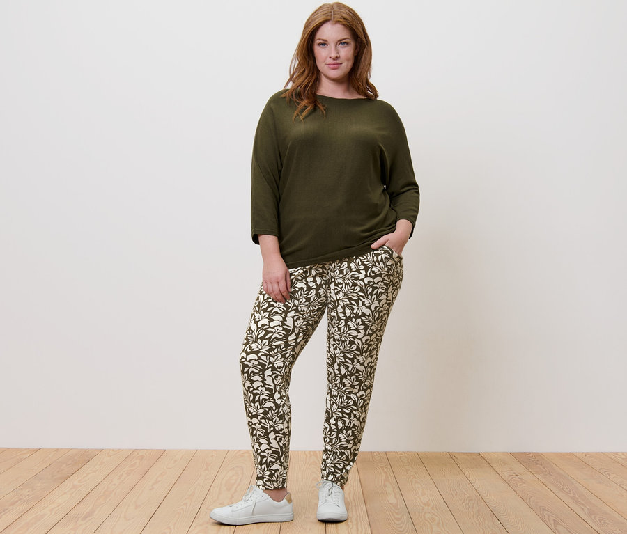 Eine Frau steht und trägt einen grünen Feinstrickpullover und eine bedruckte Jerseyhose.