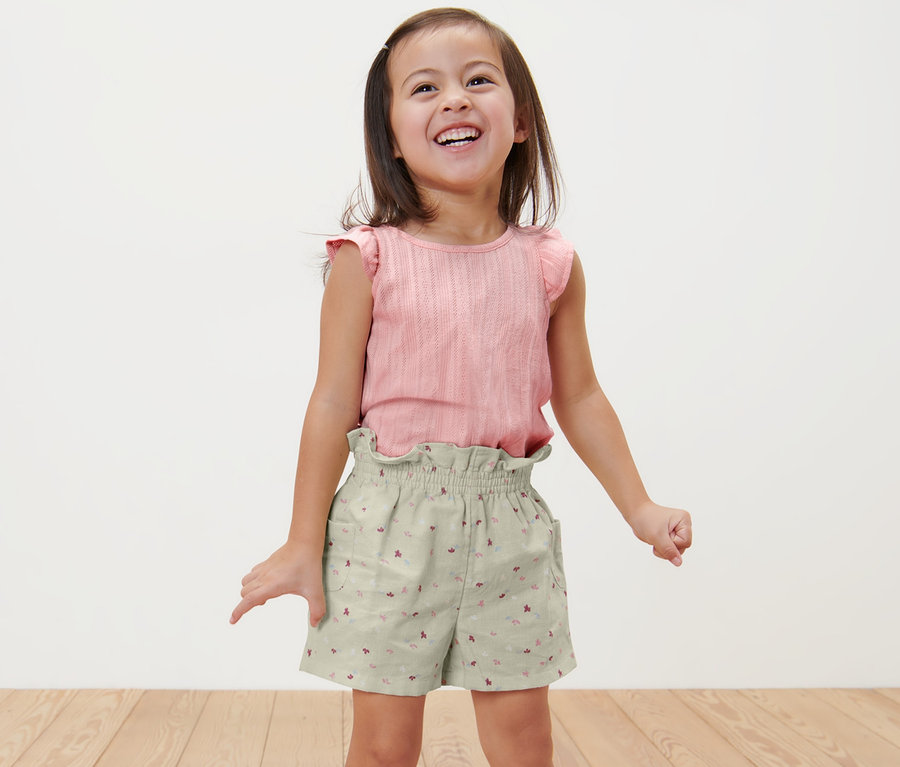 Lächelndes Mädchen steht und trägt Kinder-Paperbag-Shorts und ein rosa Oberteil.