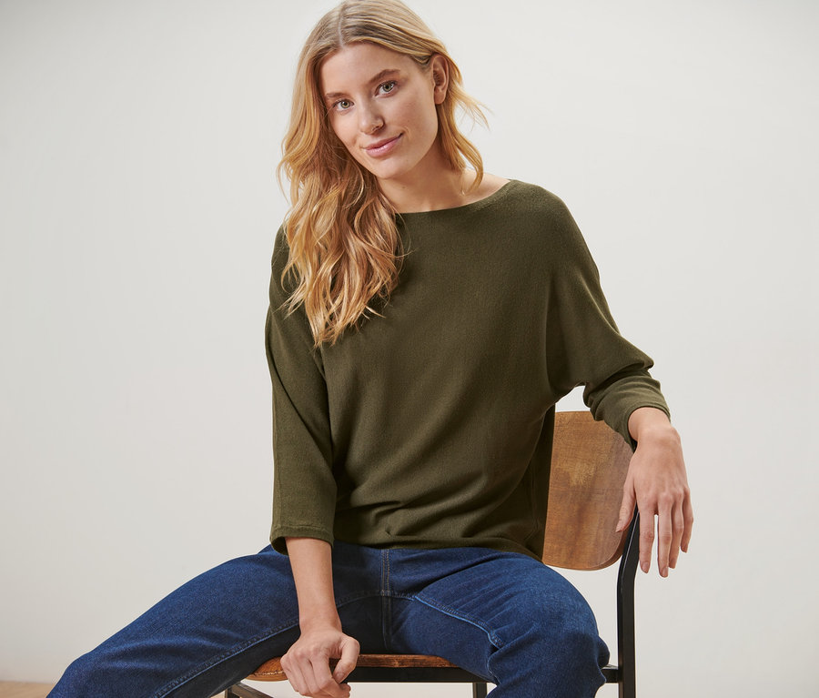 Blonde Frau sitzt auf einem Stuhl und trägt einen grünen Feinstrickpullover und blaue Jeans.