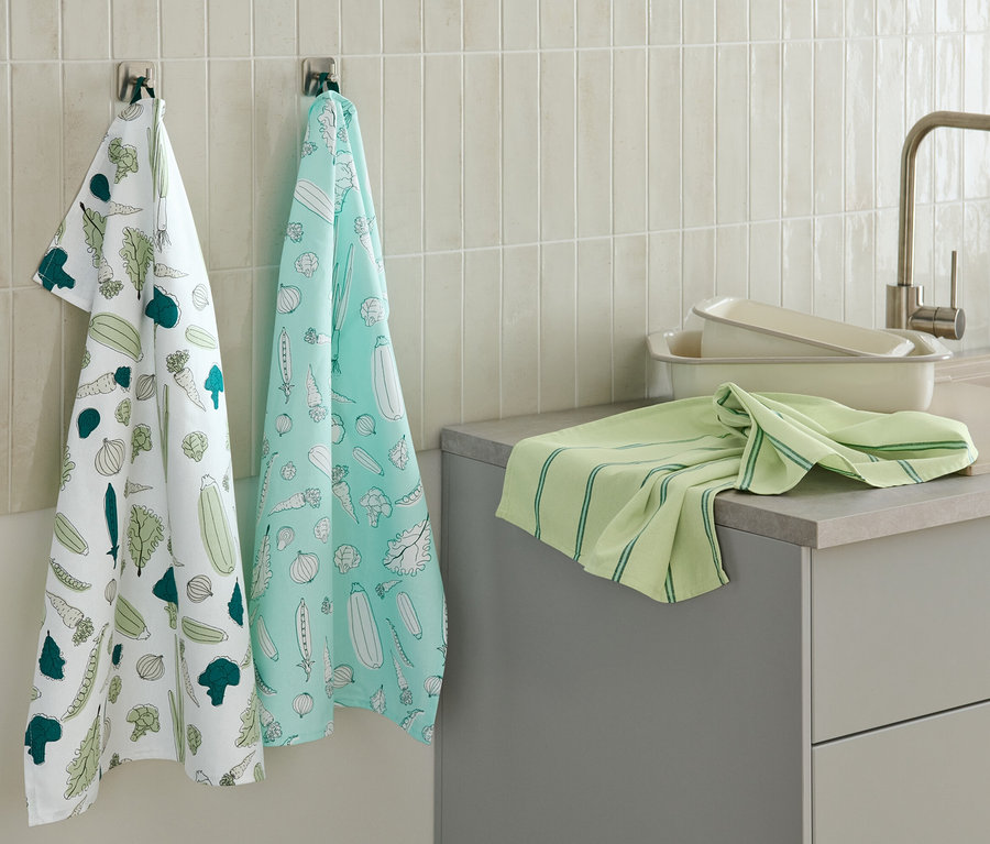 Zwei Geschirrtücher mit Gemüseaufdruck hängen an der Wand, ein weiteres grünes Geschirrtuch liegt auf der Küchentheke.