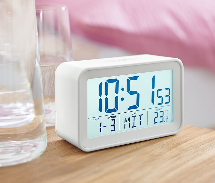Ein weißer digitaler LCD-Wecker steht auf einem Nachttisch neben einem Glas Wasser.