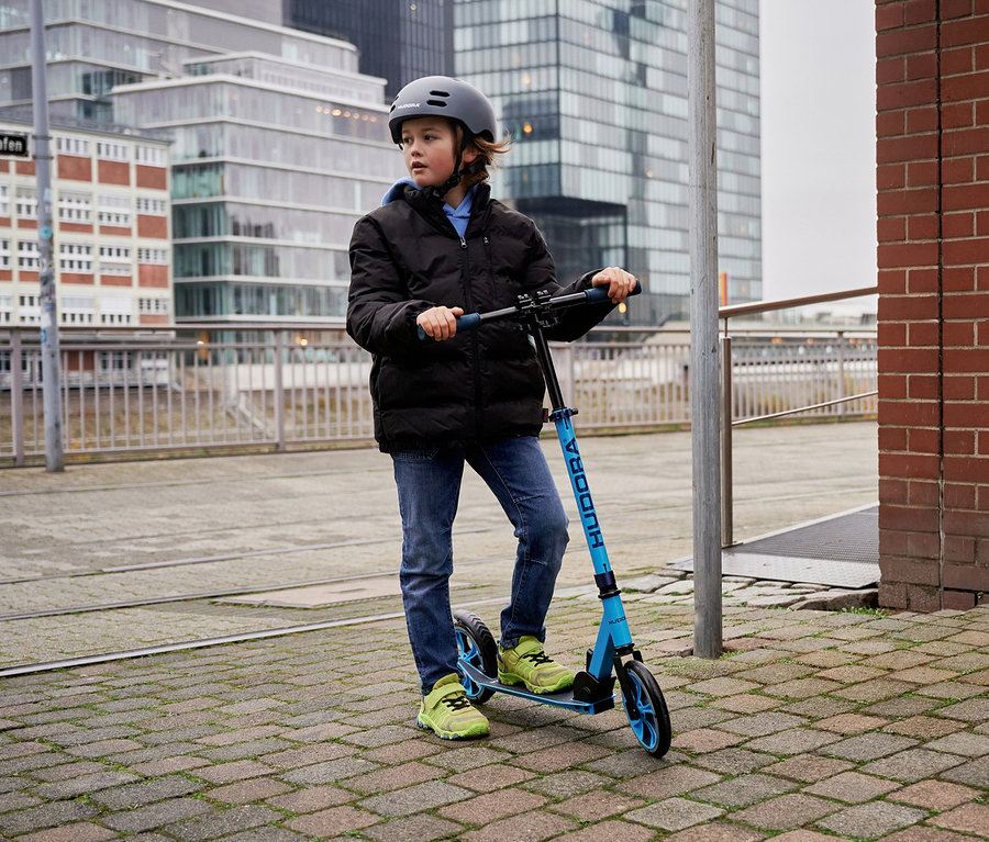 Ein Junge auf einem blauen HUDORA Scooter Up 200 mit Helm steht auf dem Gehweg.