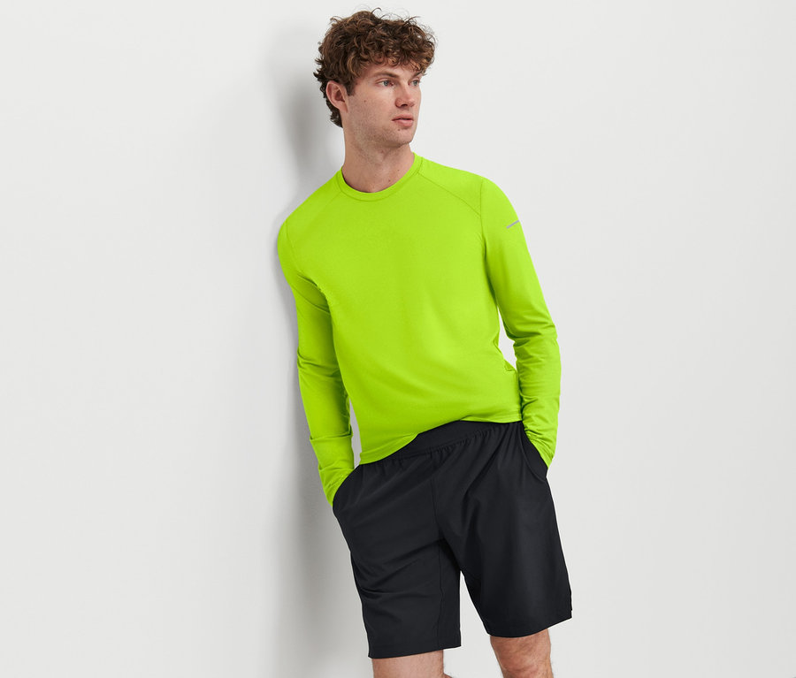Mann lehnt an Wand, trägt neon grünes Langarmshirt und Sportshorts.