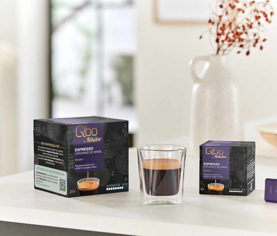 Zwei schwarz-violette Qbo Espresso CAFEZINHO DO BRASIL Kapselpackungen (27 und 8 Stück), ein Glas Espresso und eine einzelne violette Kapsel stehen auf einem weißen Tisch. Im Hintergrund steht eine weiße Vase mit roten Beeren.