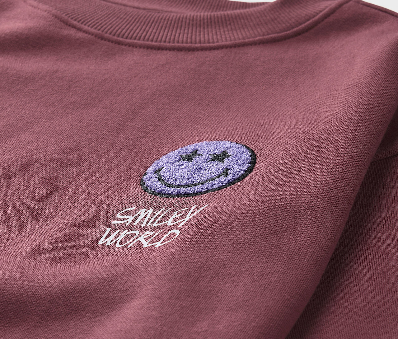 Detailansicht eines bordeauxroten Kinder-Sweatshirts SMILEYWORLD® mit angerauter Innenseite und lila Smiley-Applikation.