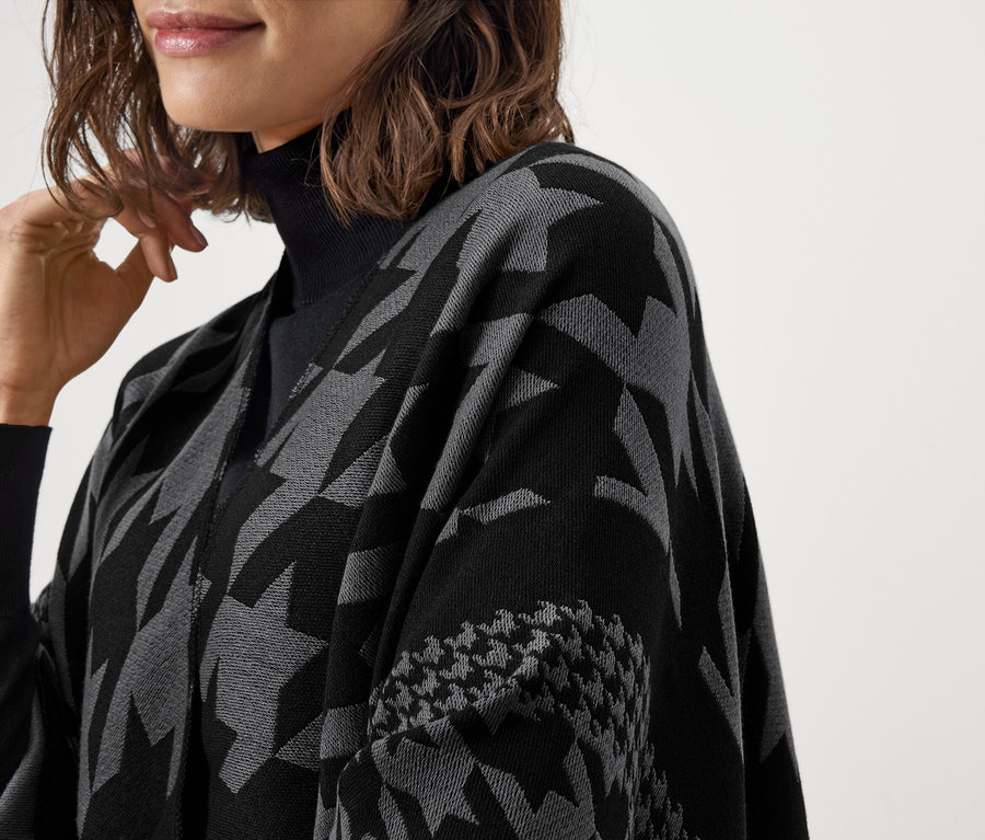 Nahaufnahme einer Frau mit schwarzem Rollkragenpullover, die eine grau-schwarze Pelerine mit Muster trägt.