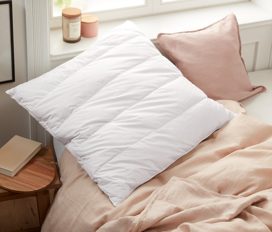 Auf einem Bett liegen ein weißes Steppkissen und ein rosafarbenes Kissen neben einem Zirbenkopfkissen.