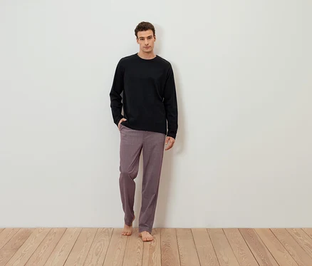 Mann steht barfuß vor einer weißen Wand, trägt ein schwarzes Langarmshirt und gemusterte Hosen.
