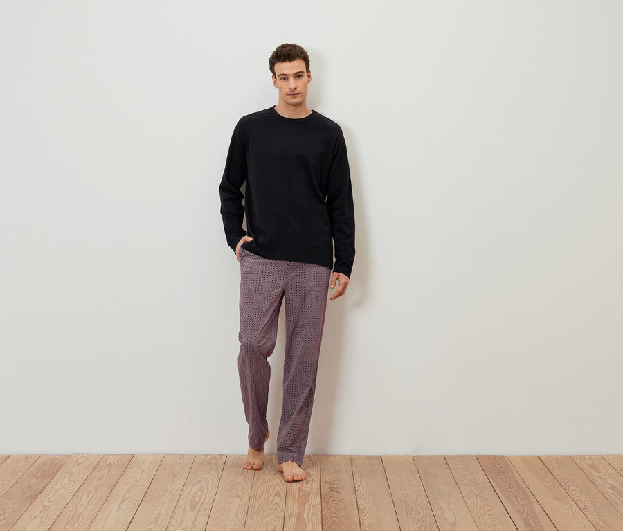 Mann steht barfuß vor einer weißen Wand, trägt ein schwarzes Langarmshirt und gemusterte Hosen.