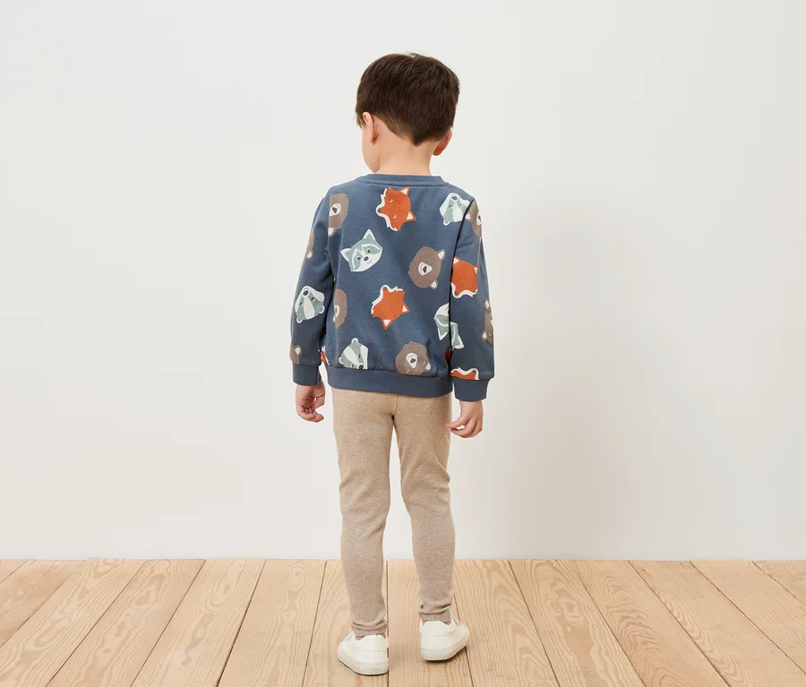 Ein Junge steht mit dem Rücken zur Kamera und trägt ein blaues Sweatshirt mit Tiermuster, beige Leggings und weiße Schuhe.