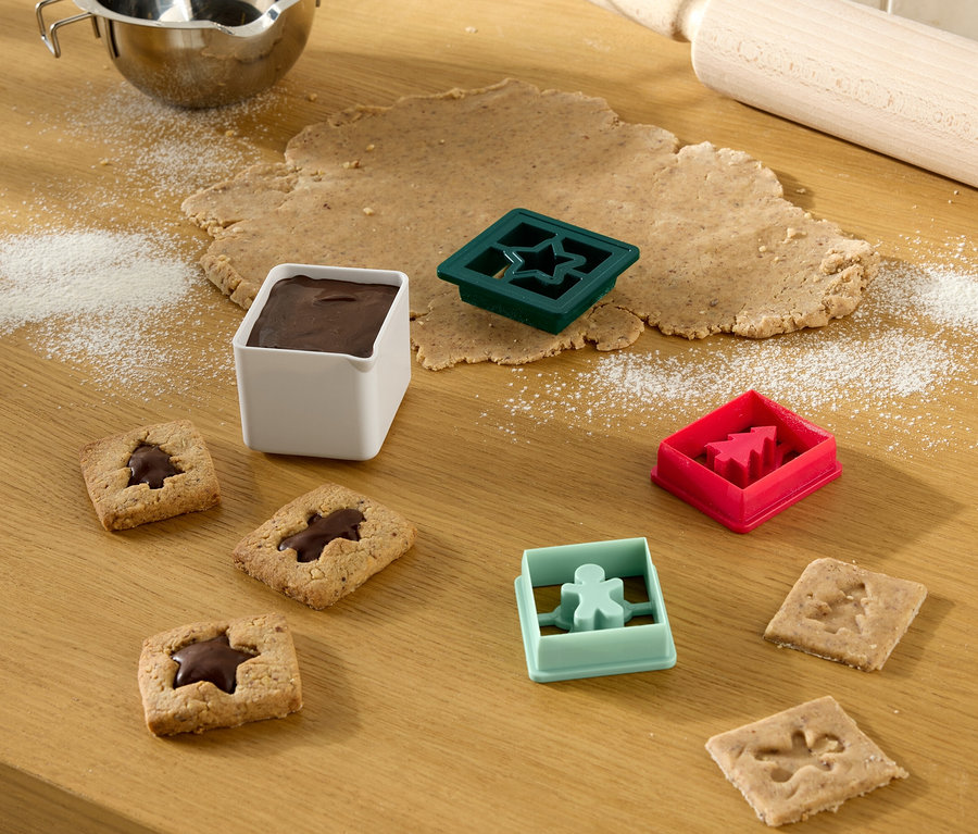 Auf einem Holztisch liegen ausgerollter Teig, Ausstechformen, fertige Kekse und eine Schüssel mit Zuckerguss.
