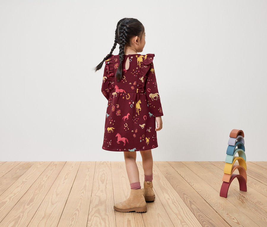 Mädchen steht mit dem Rücken, trägt ein burgunderrotes Kleid mit Pferdeaufdruck und Spielzeug.