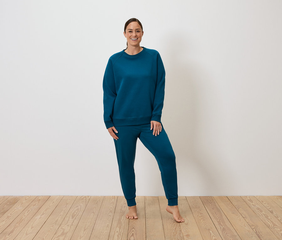 Eine Frau posiert in einem blauen Trainingsanzug auf einem Holzboden.