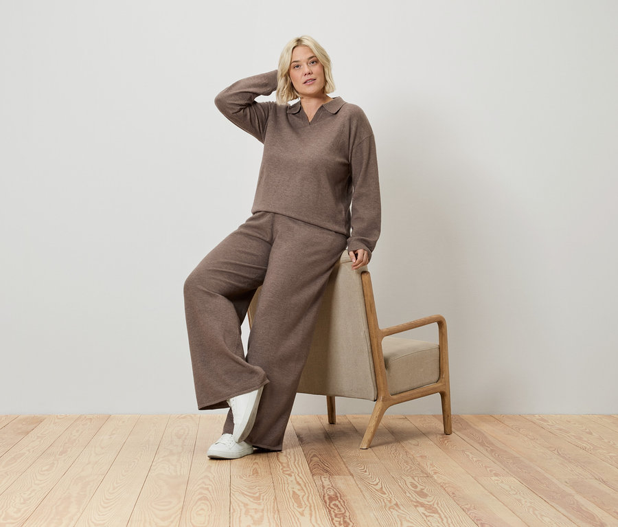 Blonde Frau in braunem Pullover und weiter Hose posiert an einen Stuhl gelehnt.