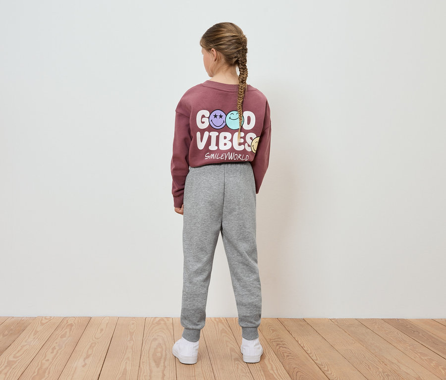Mädchen mit Zopf trägt ein weinrotes Sweatshirt mit der Aufschrift Good Vibes und eine graue Jogginghose.