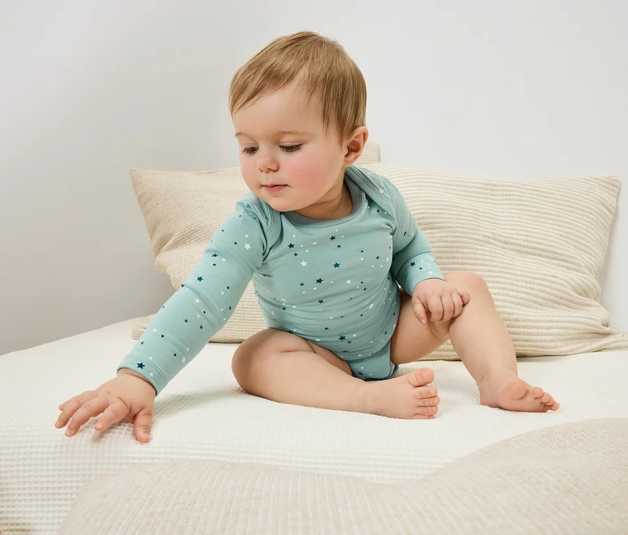 Ein Baby sitzt auf einem Bett und streckt seine Hand nach einer Decke aus.
