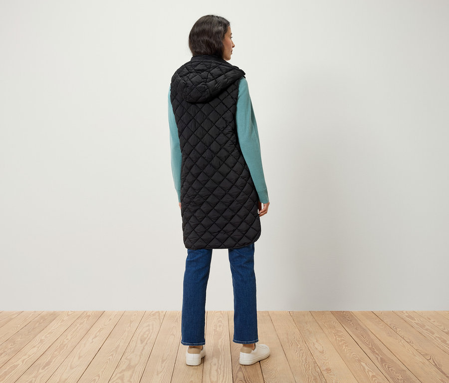 Frau steht mit dem Rücken, trägt eine lange, gesteppte, schwarze Weste mit Kapuze, einen blauen Pullover und Jeans.
