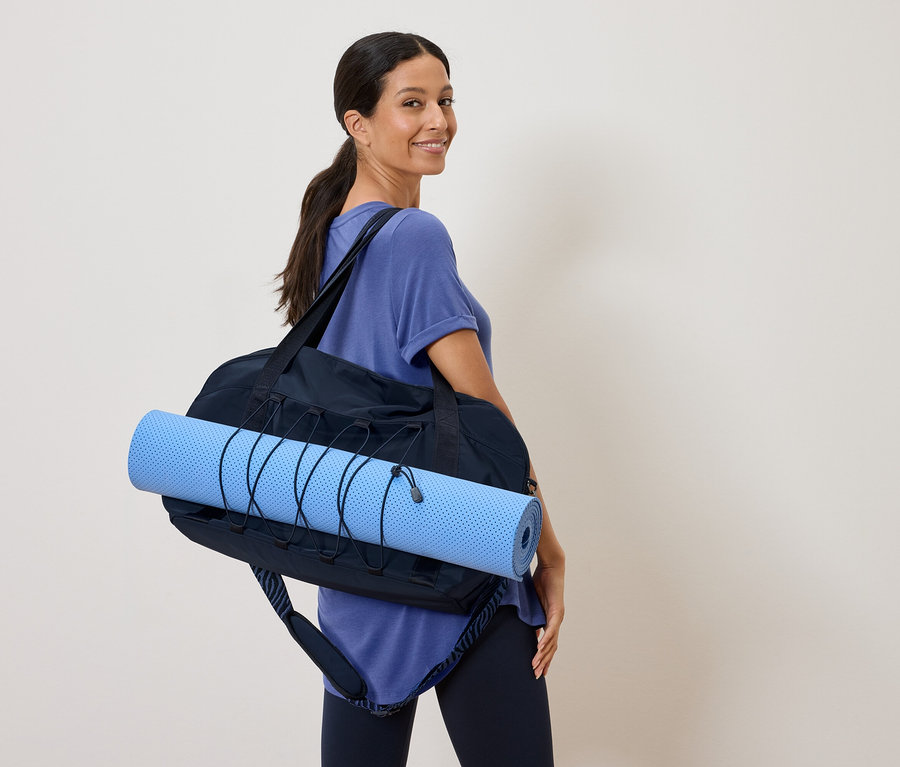 Frau trägt eine blaue Yogamatte in einer schwarzen Sporttasche über ihrer Schulter und blickt über ihre Schulter.