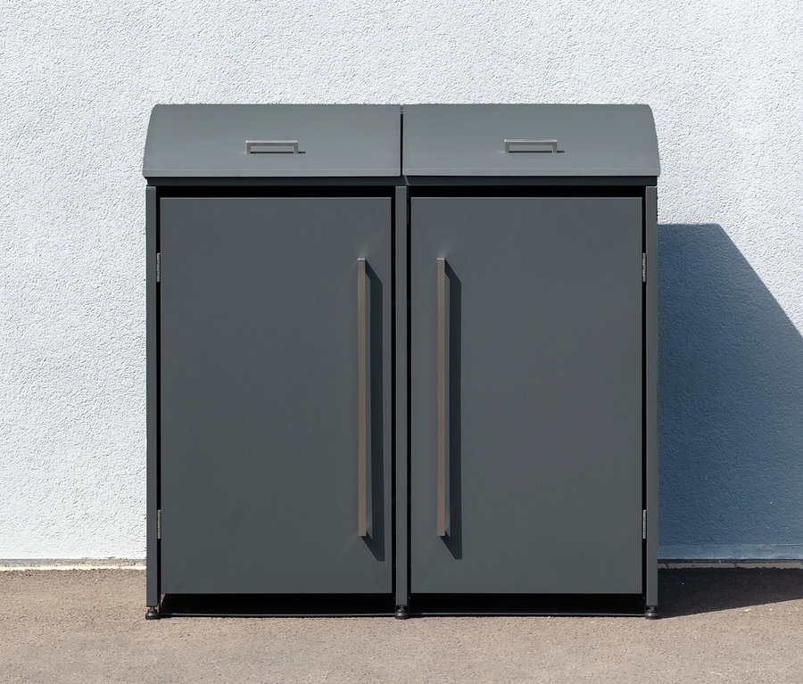 Eine graue Mülltonnenbox, 2er, steht draußen vor einer weißen Wand.