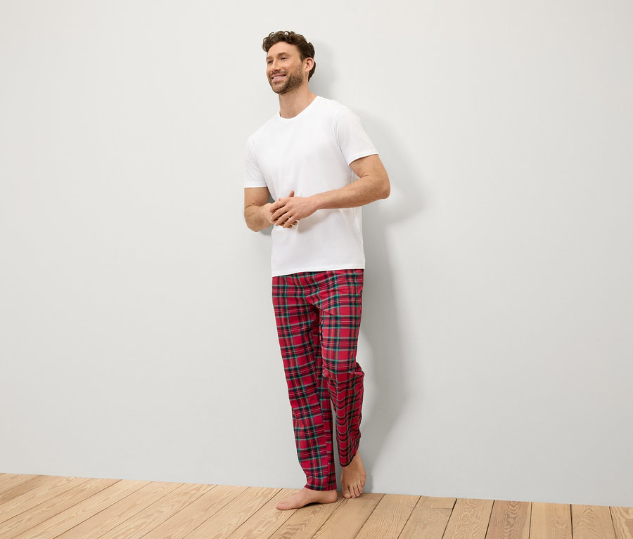 Ein lächelnder Mann steht an einer Wand und trägt ein weißes T-Shirt und eine rot karierte Hose.