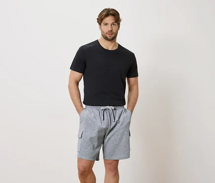 Mann mit Händen in den Taschen einer grauen Kordelzugshorts und einem schwarzen Kurzarm-T-Shirt.