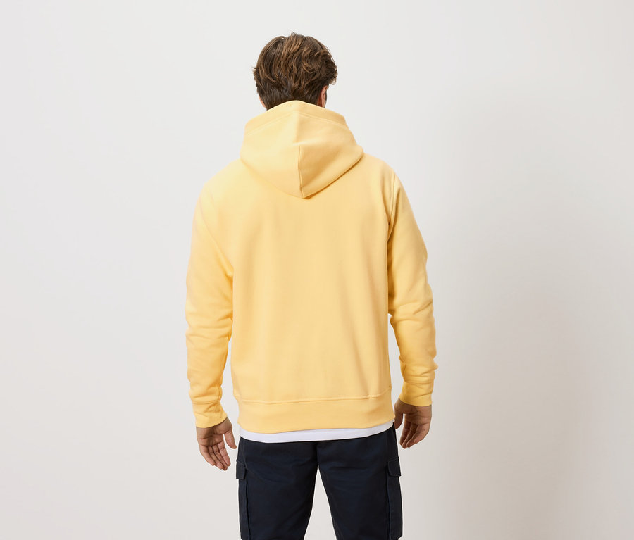 Rückansicht eines Mannes mit blonden Haaren, der einen gelben Kapuzenpullover und dunkle Hosen vor einem hellen Hintergrund trägt.