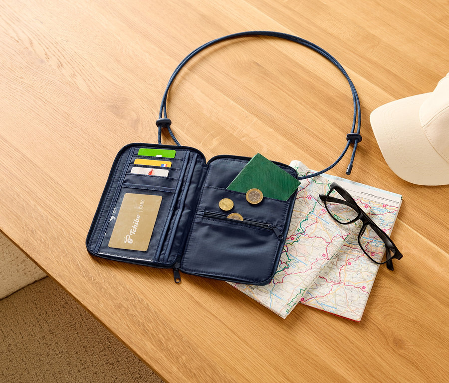 Offene, marineblaue Reisegeldbörse mit Karten, Reisepass und Münzen auf einem Holztisch, zusammen mit einer Karte, einer schwarzen Brille und einer weißen Kappe.