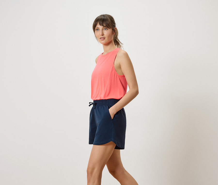 Frau mit korallfarbenem Tanktop und marineblauen Shorts, Hand in der Hosentasche, seitliche Ansicht.