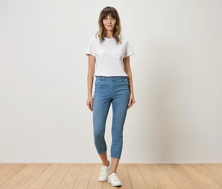 Frau mit weißem T-Shirt und blauen Jeans steht vor einer Wand.