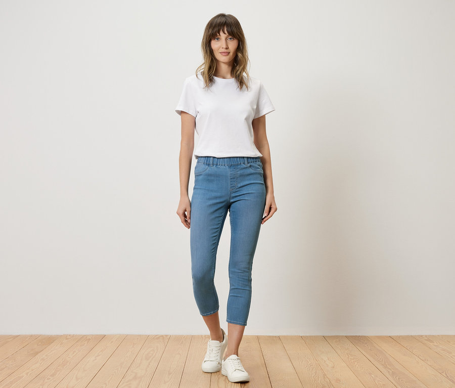 Frau mit weißem T-Shirt und blauen Jeans steht vor einer Wand.