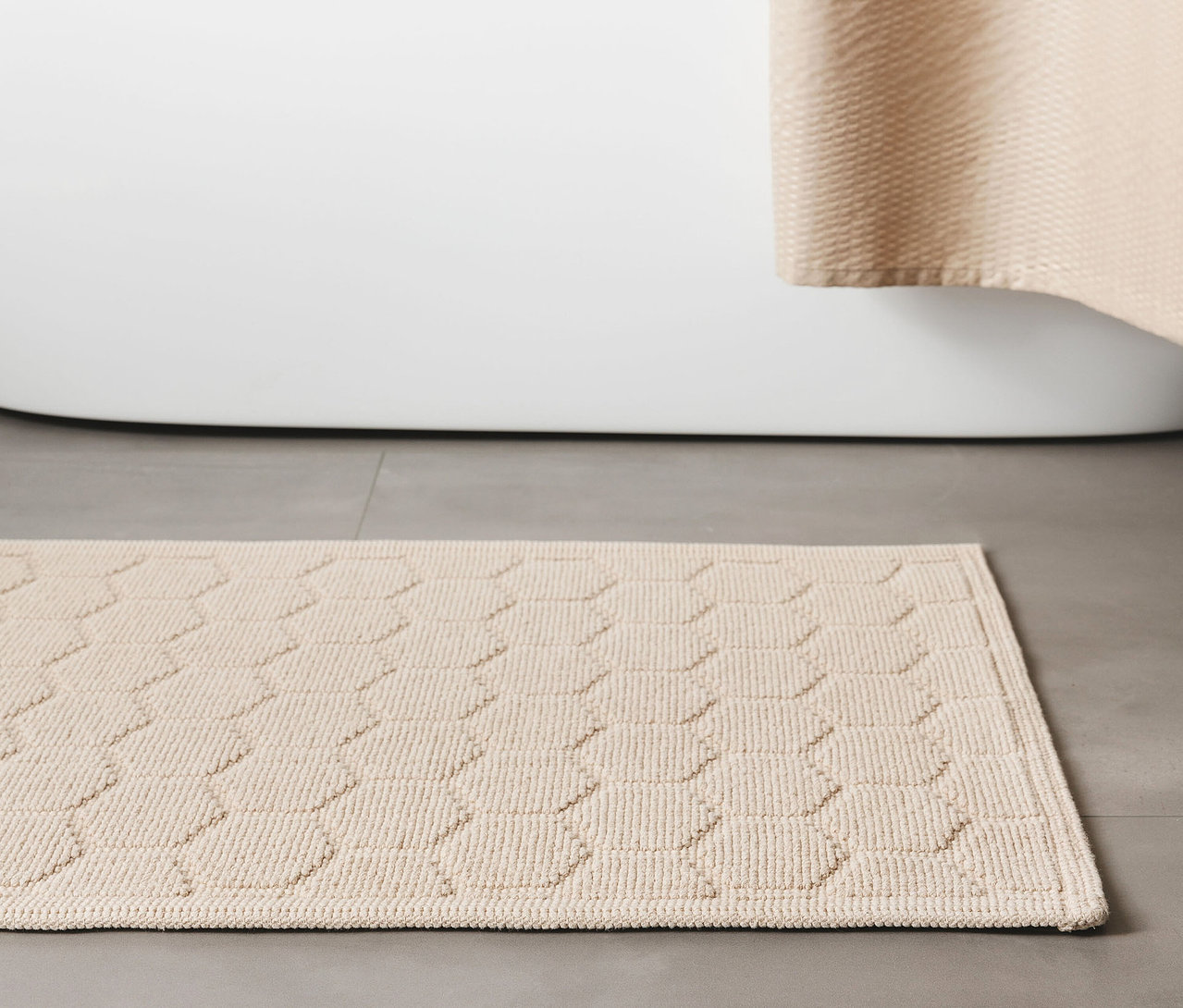 Beige Badematte »Jojo« von benuta mit Rautenmuster, 50 x 80 cm, liegt vor einer weißen Badewanne auf grauem Fliesenboden.