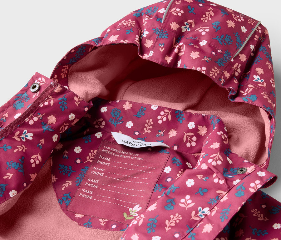 Nahaufnahme einer burgunderfarbenen Kinderjacke mit Kapuze und Blumenmuster, innen mit rosa Fleece gefüttert. Ein Etikett mit Platz für Name und Telefonnummer ist sichtbar.
