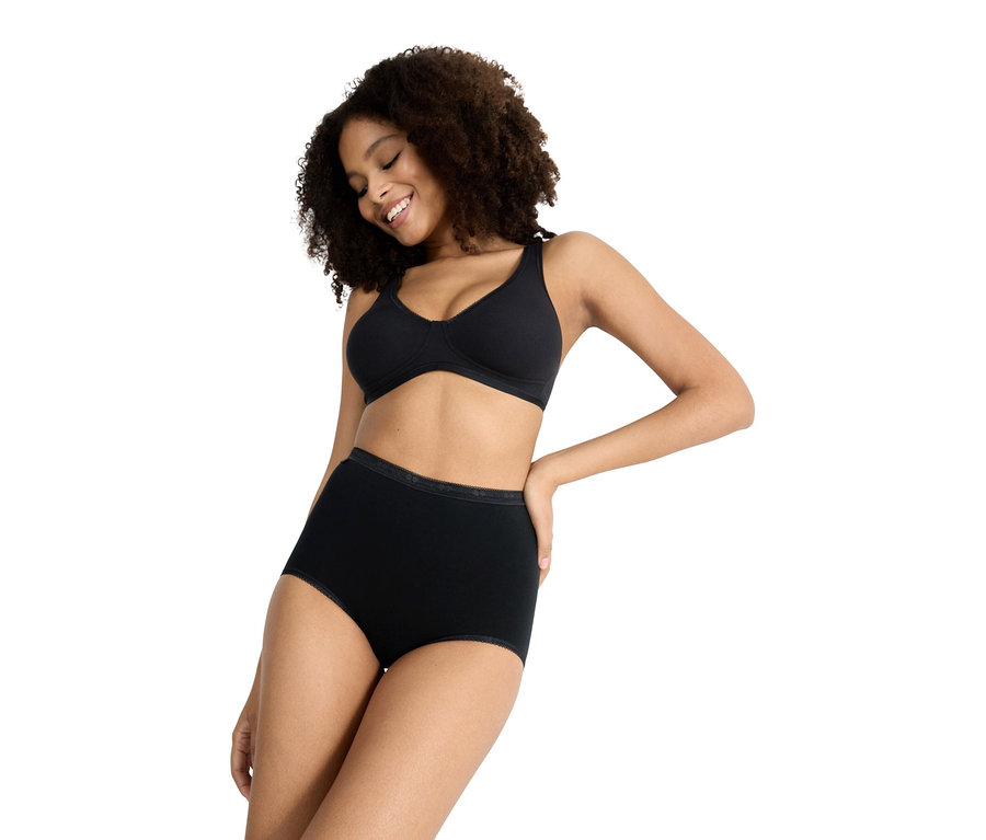 Frau mit lockigem Haar trägt einen schwarzen BH und schwarze, hoch taillierte Slips, die leicht nach unten schaut.