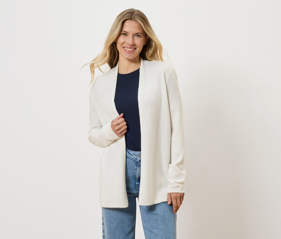 Blonde Frau trägt eine offene, weiße Strickjacke über einem marineblauen T-Shirt und blauen Jeans.