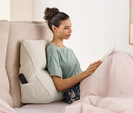 Frau sitzt im Bett und liest ein Buch, gestützt von einem Kissen mit einer Fernbedienung in der Seitentasche.