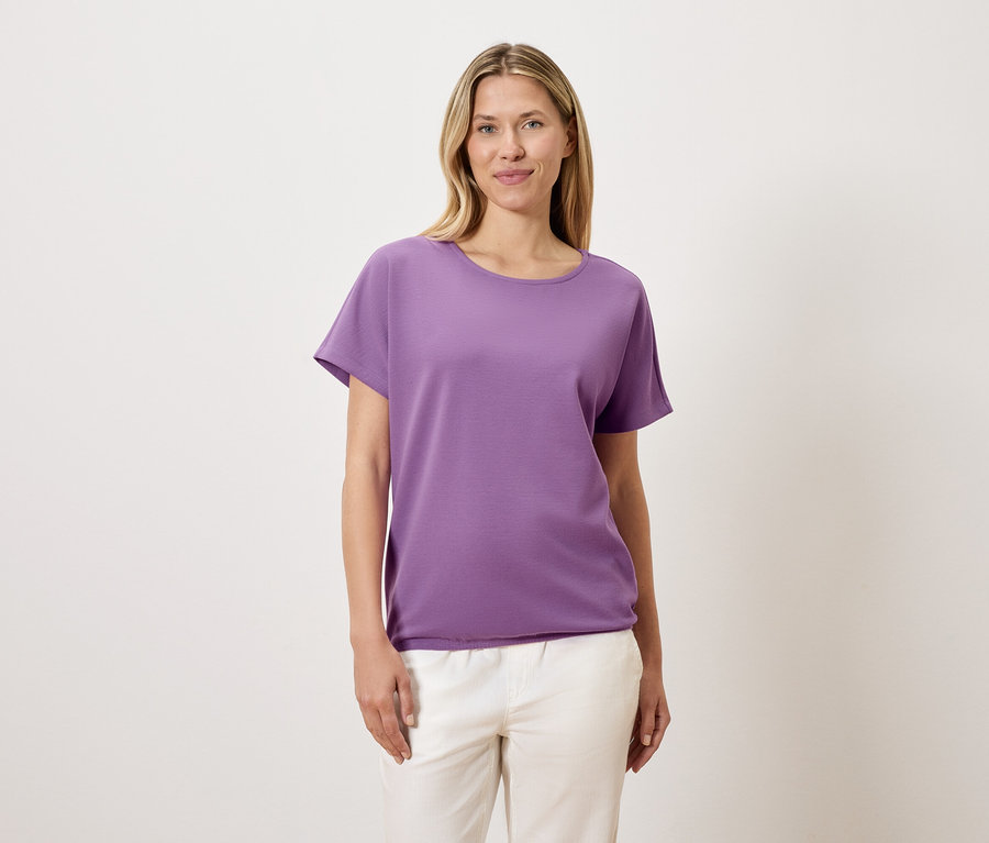Frau mit blonden Haaren trägt ein violettes T-Shirt und weiße Hosen.