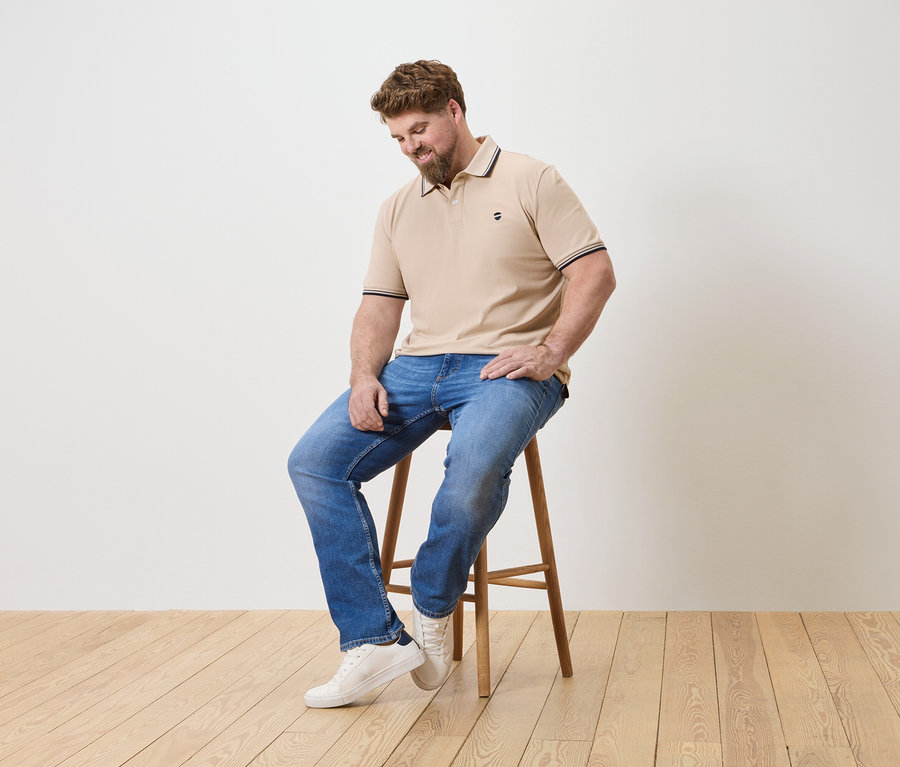 Mann mit Bart sitzt auf einem Hocker, trägt ein beiges Poloshirt, blaue Jeans und weiße Turnschuhe.