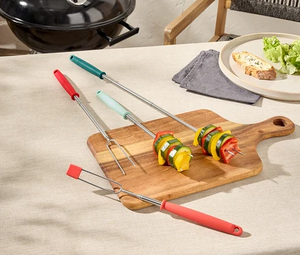 Drei Grillspieße mit Gemüse auf einem Holzbrett, ein Grill und ein Teller mit Brot im Hintergrund.