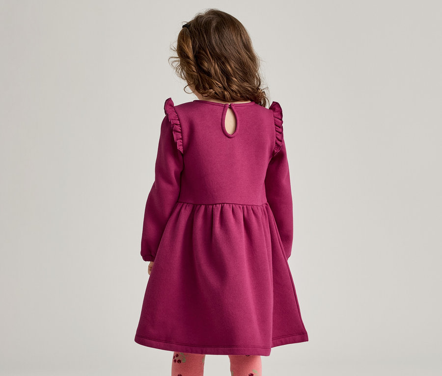 Rückansicht eines kleinen Mädchens in einem langen, weinroten Kleid mit Rüschenärmeln und gemusterten Leggings.