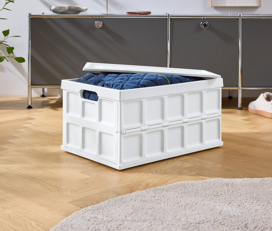 Eine weiße Faltbox mit blauem Inhalt steht auf einem Holzboden vor einem grauen Schrank und Schuhen.