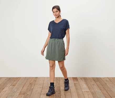 Eine Frau steht auf einem Holzboden und trägt ein marineblaues T-Shirt, einen olivgrünen Rock und marineblaue Stiefel.