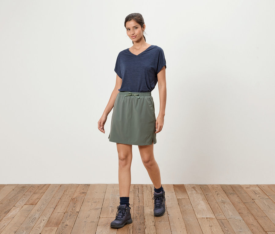 Eine Frau steht auf einem Holzboden und trägt ein marineblaues T-Shirt, einen olivgrünen Rock und marineblaue Stiefel.