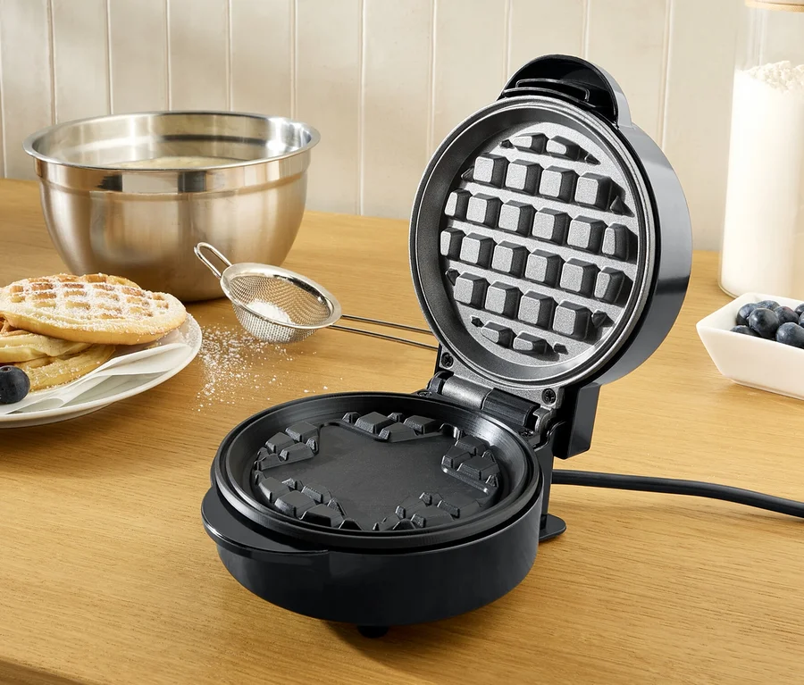 Ein offenes, schwarzes Waffeleisen auf einer hölzernen Arbeitsplatte, daneben ein Teller mit mit Puderzucker bestreuten Waffeln und Blaubeeren.