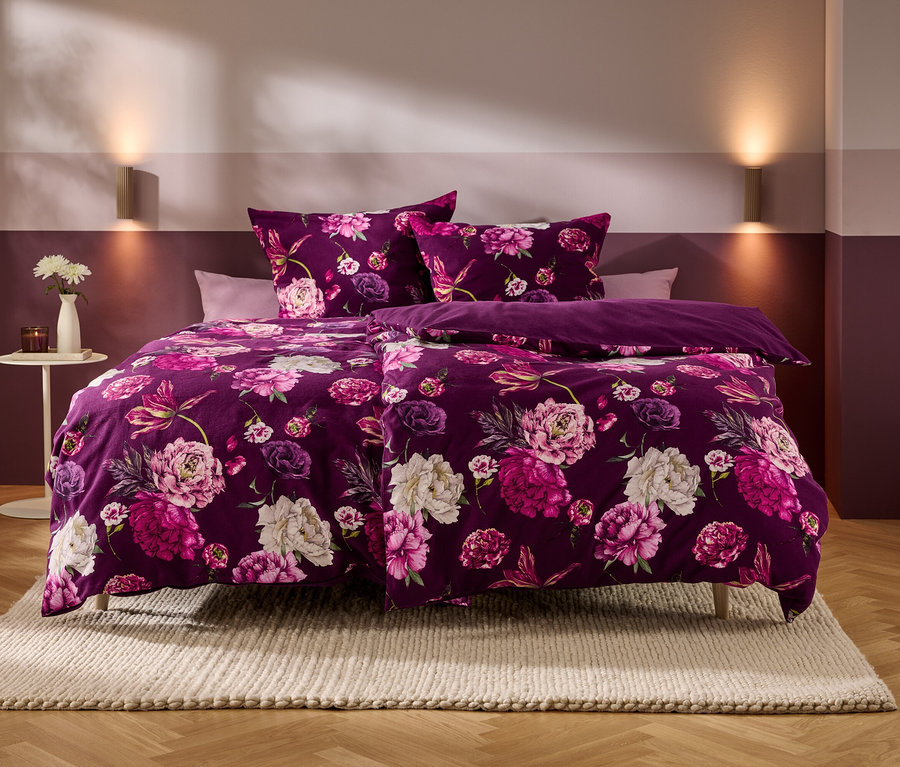 Ein Bett mit violetter Bettwäsche, die ein Muster aus rosa und weißen Blumen zeigt, steht in einem Schlafzimmer mit zweifarbigen Wänden und Holzboden. Auf einem Beistelltisch stehen eine Vase mit weißen Blumen und eine Kerze.