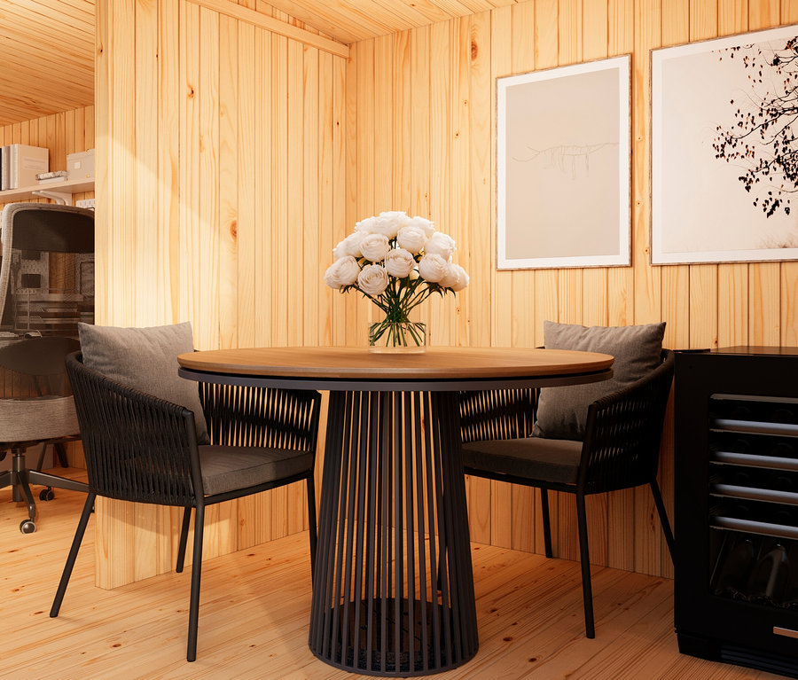 Ein Raum mit Holzwänden und -boden, in dem ein runder Holztisch mit schwarzem Fuß und zwei Stühlen mit grauen Kissen stehen. Auf dem Tisch steht eine Vase mit weißen Blumen. Im Hintergrund sind zwei gerahmte Bilder und ein Bürobereich zu sehen.