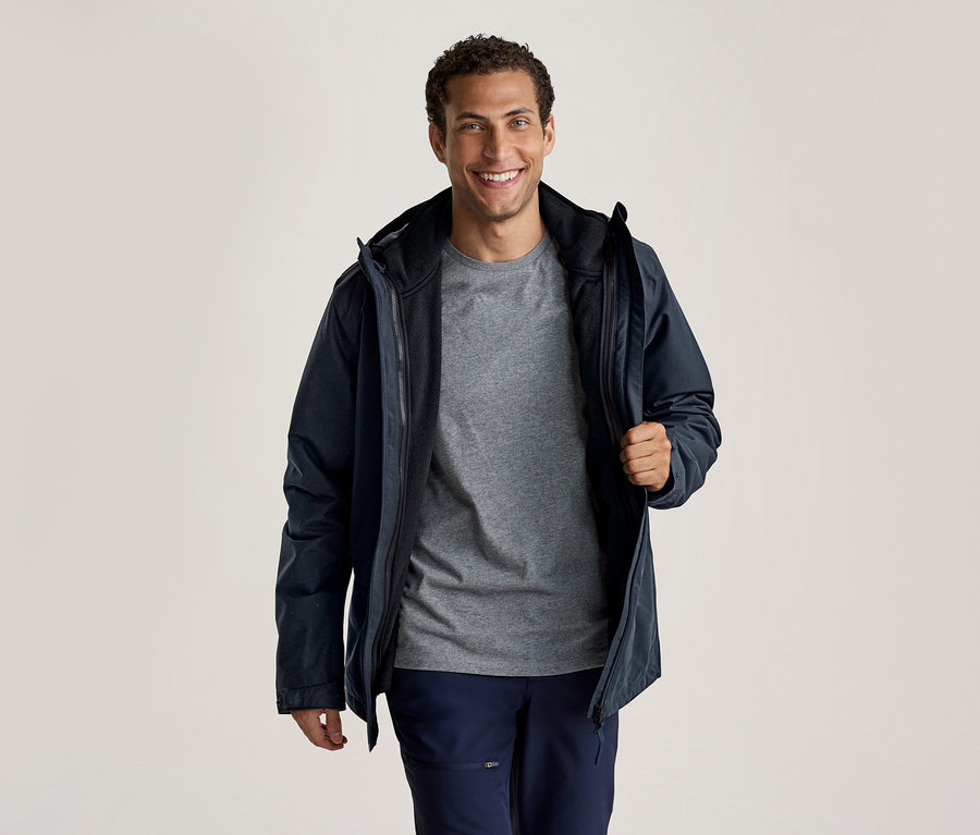 Ein lächelnder Mann trägt eine dunkelblaue Jacke über einem grauen T-Shirt und dunkelblauen Hosen. Er hält die Jacke mit der rechten Hand offen.